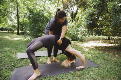 Adult trainer helping unrecognizable female standing in urdhva dhanurasana pose while practicing yoga in summer park