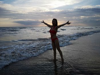 Full length of woman with arms outstretched standing on shore at beach against sky during sunset