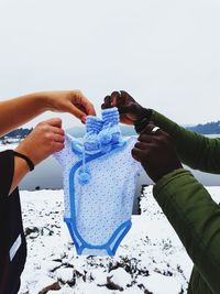 Cropped hands of couple holding baby clothing against lake and sky during winter