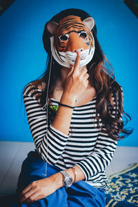 Portrait of woman wearing tiger mask while sitting against blue wall