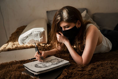 Woman wearing mask drawing in book on bed