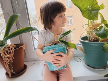 Toddler girl sitting on the window holding flower pots with plants