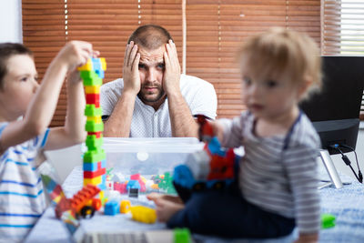 Portrait of father with head in hand while kids playing with toy on table at home