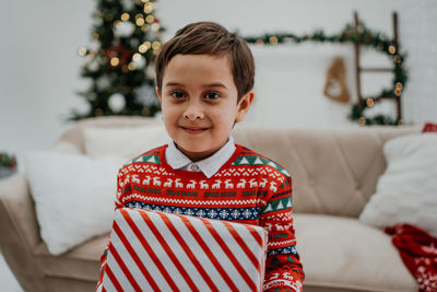 Cute little boy holding gift box and posing to the camera in the living room before the christmas