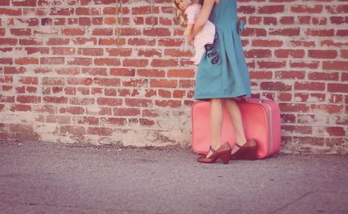 Low section of girl carrying doll and briefcase against brick wall