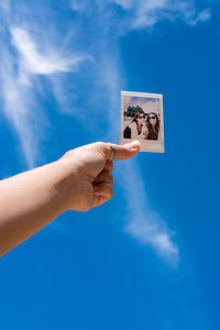 Cropped hand of person holding photograph against blue sky during sunny day