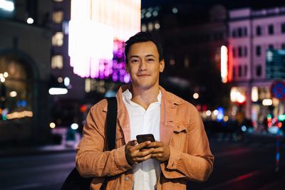 Portrait of smiling man using mobile phone while standing in illuminated city at night
