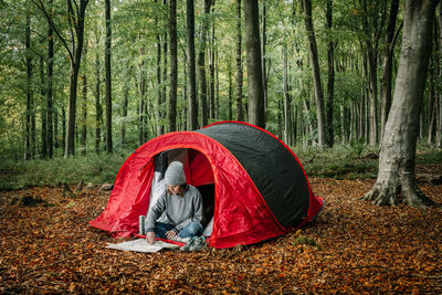 Traveling female sitting near camping tent and navigating with paper map while searching for trail during vacation in forest