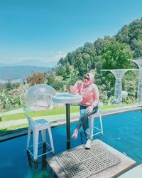 Man sitting on chair by swimming pool against sky