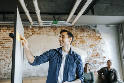 Smiling male tech professional sticking adhesive notes on whiteboard in office