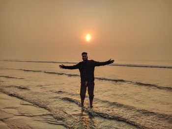 Full length portrait of man with arms outstretched standing at beach during sunset