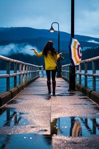 Rear view of woman with umbrella walking on pier over lake against mountains