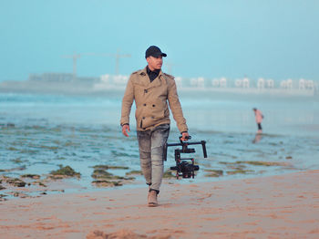 Full length of man holding camera while walking at beach