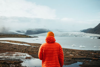 Back view of young tourist on peak of mountain in snow looking at water in valley