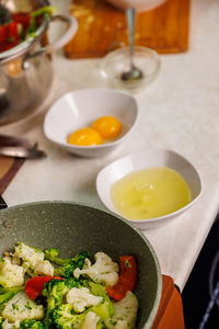 Kitchen table with bowls with separated eggs and skillet with frying vegetables in the foreground