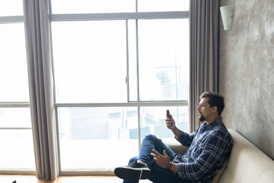 Side view of young man looking through window with smartphone 