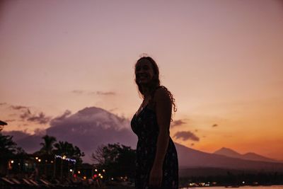 Smiling woman standing by mountains against sky during sunset