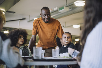 Smiling teacher standing by students during lunch break in cafeteria