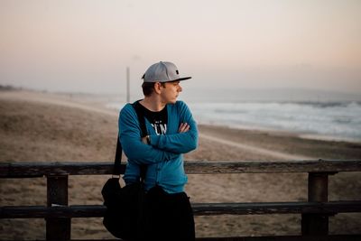 Thoughtful man with arms crossed standing on pier at beach during sunset