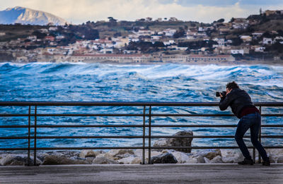 Man photographing while standing on railing by sea against sky