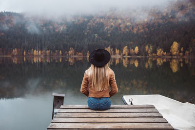 Rear view of blonde woman wearing brown jacket and hat sitting on a wooden pier  in autumn season