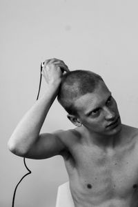 Shirtless young man shaving head with razor while looking away against wall