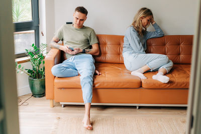 A quarreled couple is sitting on the couch, the man is using a mobile phone, the woman is offended.