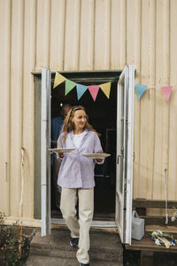 Smiling woman with food plates walking outside door during dinner party at cafe