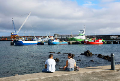 People sitting on boats moored at harbor against sky