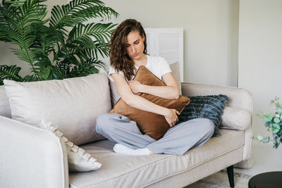 A sad woman sits on a sofa at home hugging a pillow.