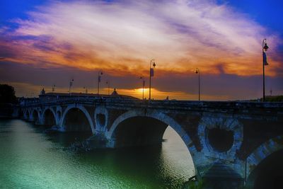 Arch bridge over calm river against cloudy sky during sunset 