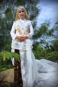 Portrait of smiling young woman standing in forest wearing traditional indonesian wedding dress.