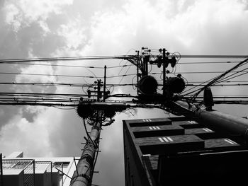 Low angle view of electricity pylons and buildings against cloudy sky