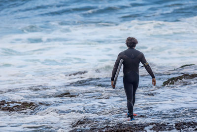 Rear view full length of man in wetsuit holding surfboard at sea