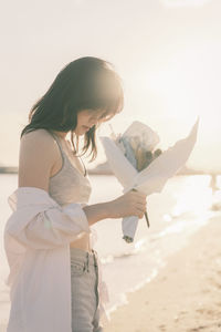 Side view of young woman with arms outstretched standing at beach holding flower