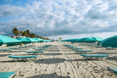 Empty deck chairs and parasols on sand at beach against cloudy sky