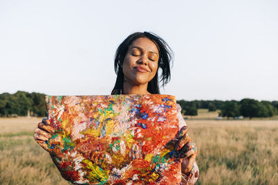 Smiling woman with eyes closed holding painted paper at park during sunset