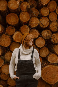 Smiling young female in warm clothes and hat looking away pensively while leaning back against heap of wooden logs stacked together in countryside