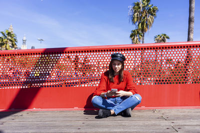 Teenage girl using mobile phone while wearing red top in city against railing during sunny day