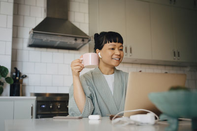 Smiling woman having coffee while on video call through laptop in kitchen