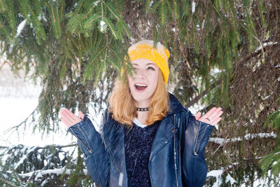 Young smiling woman in a yellow knitting hat under the fir tree at the moment of snowfall.