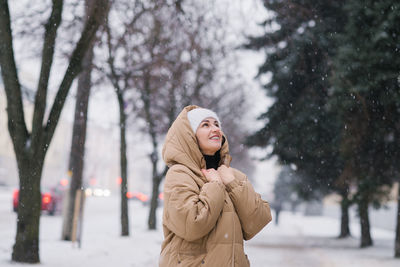 A happy woman in a hat is warming herself in a coat. it stands in the city in winter under the snow