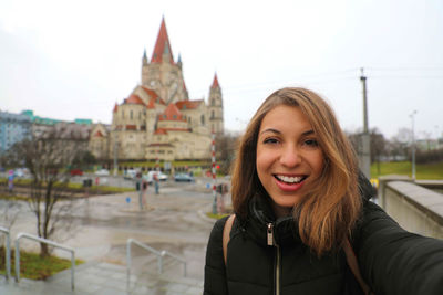 Portrait of smiling young woman standing in city against sky