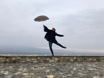 Full length of young woman jumping with umbrella on retaining wall against sky
