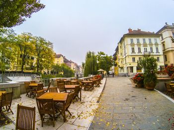 Empty chairs and tables by buildings in city against sky