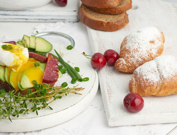 Toast with poached egg and avocado on a round board, next to croissants and ripe red 