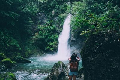 Rear view of woman standing on rock against waterfall in forest