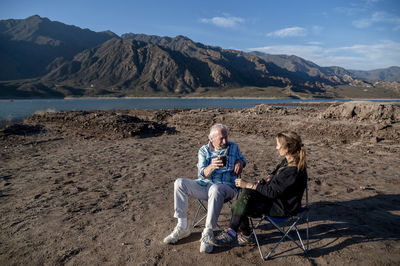 Woman and her grandfather talking while sitting on camping chairs outdoors in nature.