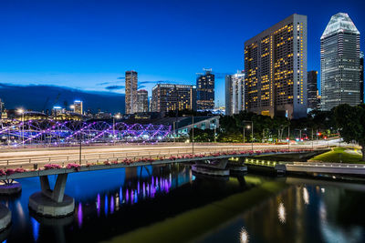 Illuminated buildings by river against blue sky at night