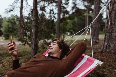 Smiling young man using mobile phone while relaxing in hammock against trees